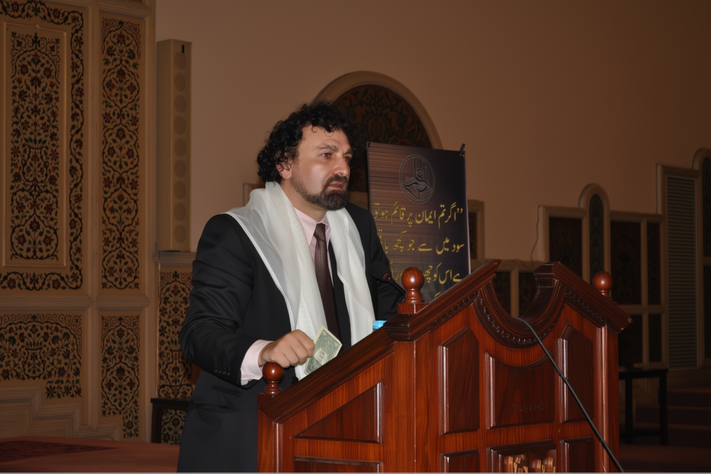 Shaykh Umar speaking at a wooden podium during a conference in a decorated hall